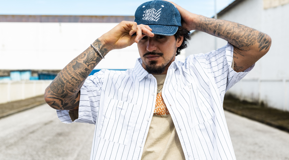 Man wearing a cap and striped shirt with tattoos on arms, standing outdoors.